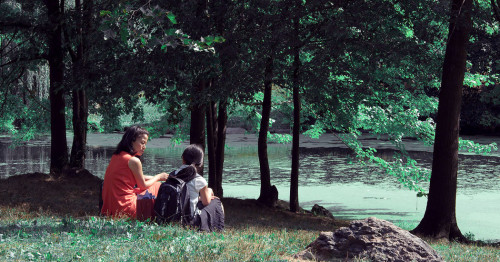 Gwen and Jules talking in the park, surrounded by green grass, tall trees, and the river.